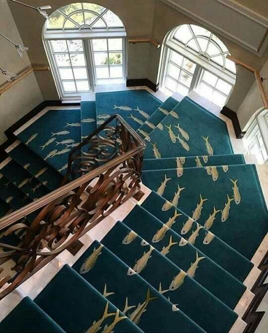 An ornate staircase with teal carpet runners featuring a pattern of yellow and white fish, viewed from above with a decorative metal railing and arched windows in the background.