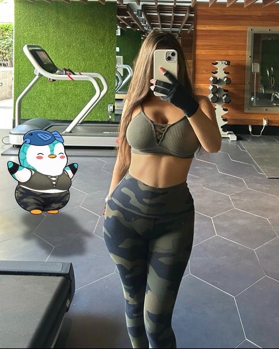 A woman takes a gym mirror selfie in athletic wear (olive sports bra and camo leggings), with a cartoon penguin sticker overlaid on the left side.