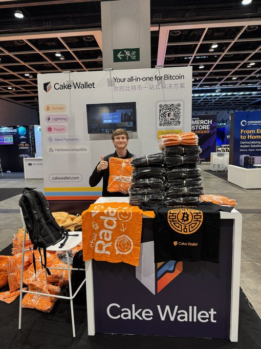 A man standing at a Cake Wallet booth at a convention, surrounded by stacked merchandise including t-shirts, hoodies, and bags, with the slogan 'Your all-in-one for Bitcoin 你的比特币一站式解决方案'.