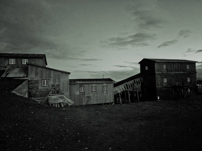 A black and white photograph of wooden mining buildings and structures under a cloudy sky, evoking a vintage industrial scene.
