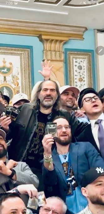 A crowd of people gather indoors in an ornate room, with a long-haired man in the center holding up a card while others take photos with their phones.
