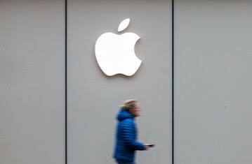A blurry photograph of a person in a blue jacket walking past a large white Apple logo on a gray paneled building exterior.