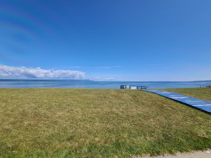 A scenic landscape featuring a grassy field leading to a calm body of water under a bright blue sky with scattered clouds, with a long blue banner laid out on the grass.