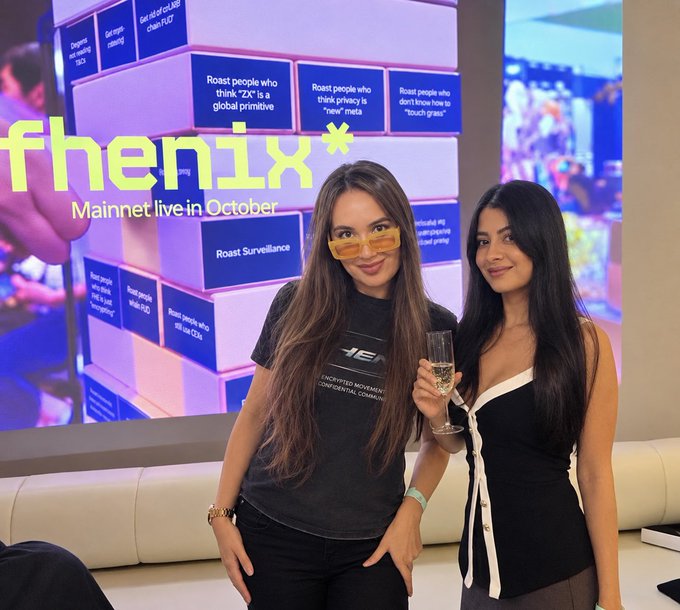 Two women pose in front of a screen displaying 'fhenix Mainnet live in October' alongside various text boxes with humorous roasts about crypto privacy.