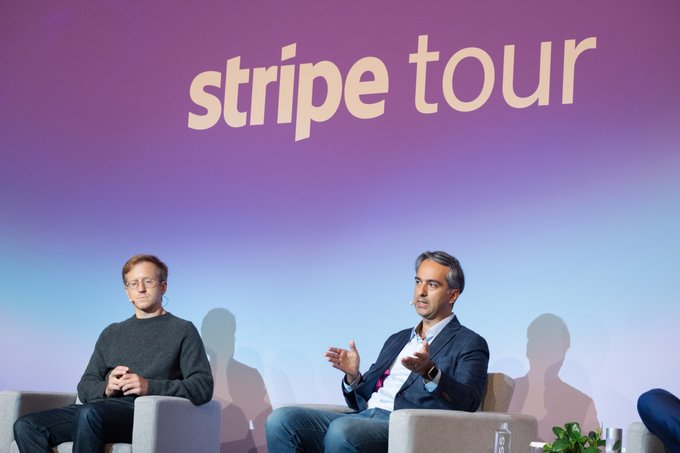 Two men sit on stage chairs in front of a purple and blue backdrop displaying 'stripe tour' in white lowercase text.