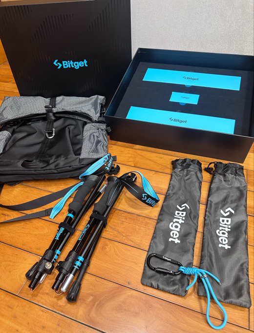 A Bitget 8th anniversary hiking gift package is displayed on a wooden floor, featuring a black backpack with teal straps, foldable hiking poles, drawstring storage bags, and a carabiner, all branded with the "Bitget" logo. The accompanying note expresses gratitude for the thoughtful gift, highlighting how the company's care for physical well-being feels especially meaningful.