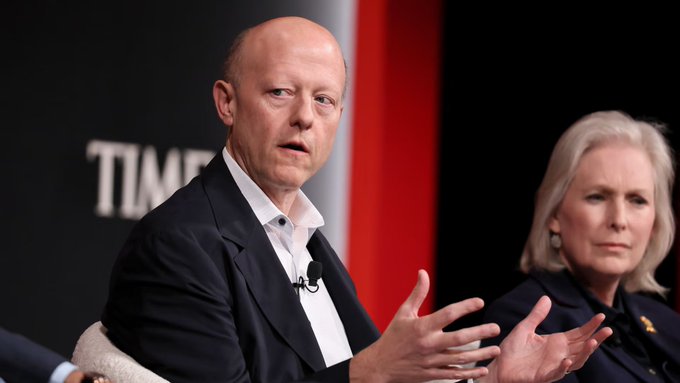 A man in a dark suit speaking with gesturing hands at a 'TIME' branded event, with an older woman with white hair seated beside him.