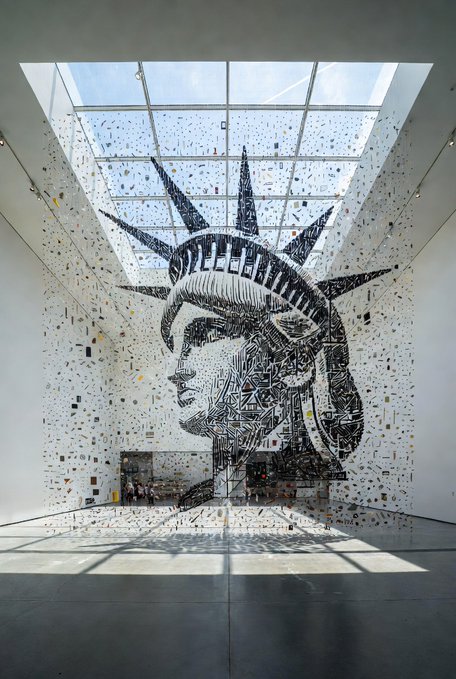 A large glass sculpture of the Statue of Liberty is suspended within a white gallery space beneath a skylight.