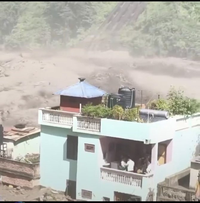 A partially obscured teal two-story house with rooftop planters sits precariously near massive flooding that has triggered a landslide or debris flow on the forested hillside behind it, endangering the residents visible on the balcony. The scene powerfully illustrates the harrowing reality of being trapped as floodwaters and landslides close in, evoking the helplessness of simply watching and waiting for rescue.