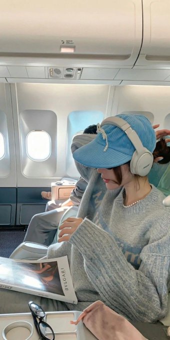 A woman wearing a blue cap and gray sweater sits on an airplane reading a Bvlgari magazine while wearing white headphones, sharing a casual "gm" (good morning) travel moment.