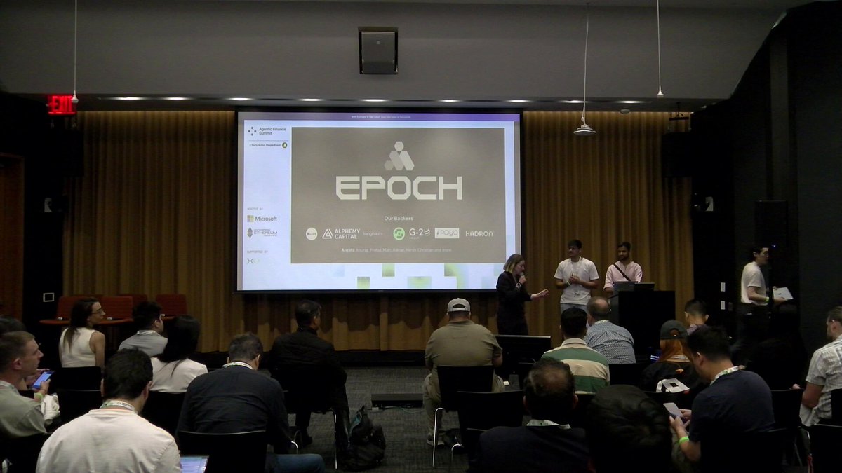 The image shows an audience of people sitting in chairs around a large screen displaying a presentation about Epoch. The attendees are engaged with the content being presented by two speakers who are standing behind the screen, likely discussing the various aspects of the platform or product they are showcasing. The room is filled with people, and some individuals have their cell phones out, possibly taking notes or capturing photos related to the event.
