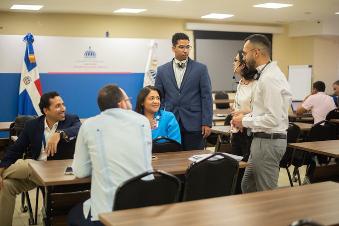 A group of people converse around a table in a classroom setting, with a Dominican Republic flag and another flag visible in the background, suggesting a formal meeting or educational seminar.