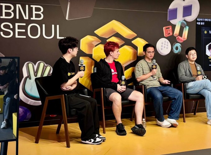 Four men sit on chairs on a stage at a 'BNB SEOUL' event, holding microphones, with colorful BNB crypto-themed decorations behind them.