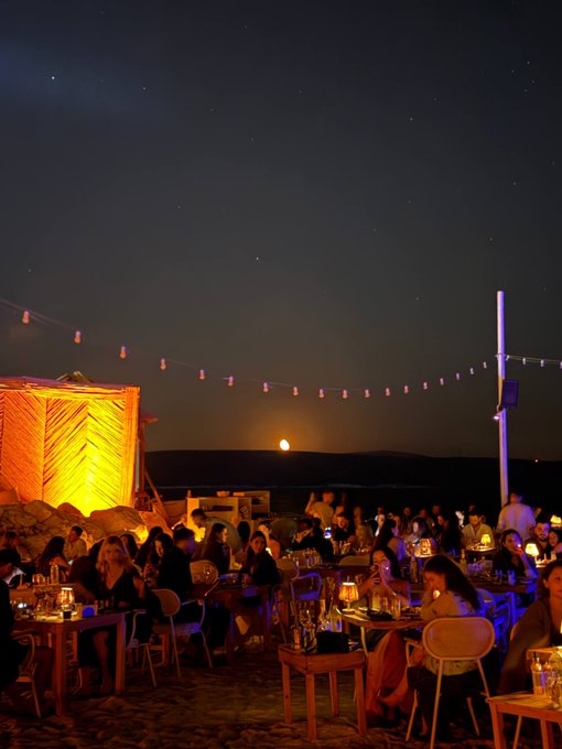 A nighttime outdoor beachside restaurant scene features string lights overhead, patrons seated at candlelit tables, and a full moon rising over the water.