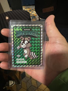 A hand holding a holographic trading card featuring a raccoon character, with the text 'SUPER STICKERS' and 'BRAVEST RACOON' visible on the card.