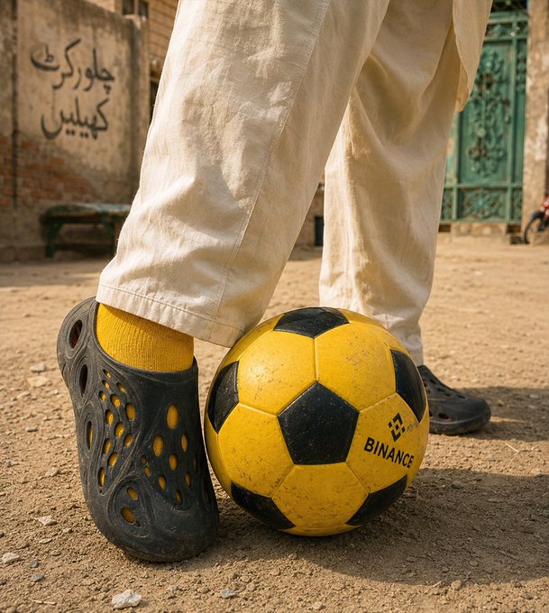 Close-up photo of a person wearing beige pants and dark crocs standing next to a worn yellow and black soccer ball branded with the 'BINANCE' logo on a dirt surface.