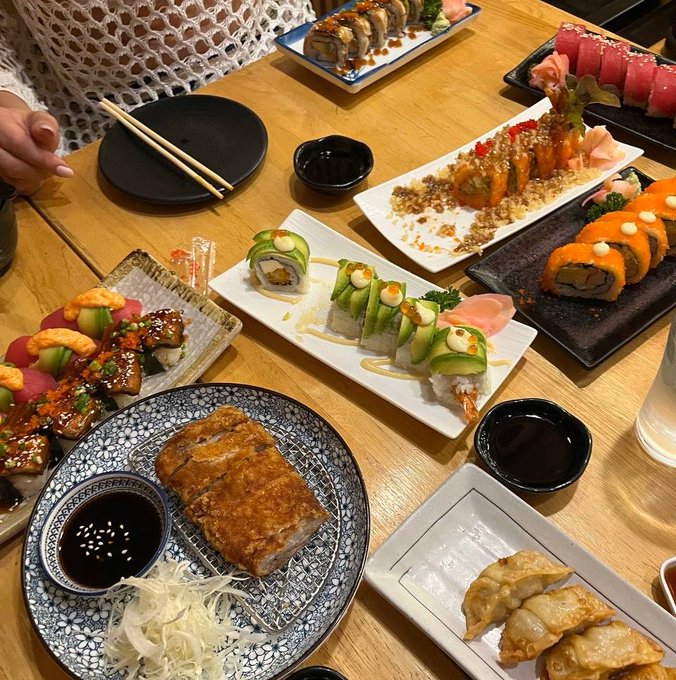 A spread of Japanese cuisine including assorted sushi rolls, tempura, dumplings, and grilled eggplant is served across multiple plates on a wooden table, paired with a caption highlighting the affordable 0.5 sol price in Thailand—suggesting an impressive value for such a diverse and abundant meal.