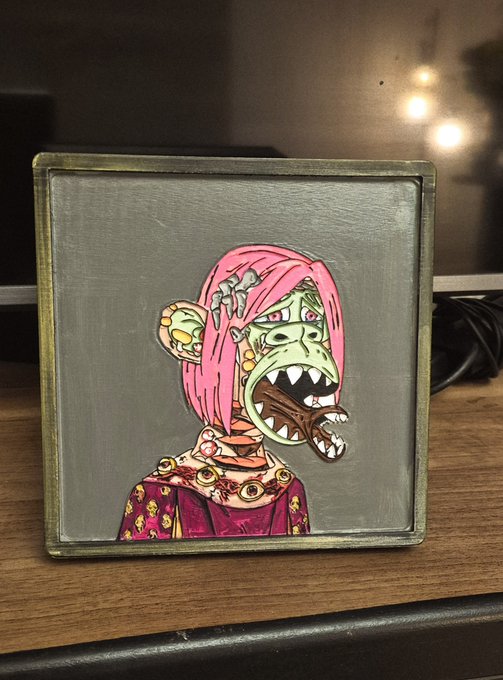A framed mixed-media artwork featuring a character with pink hair, a green monstrous face with sharp teeth, and decorative jewels on a silver/gray background.