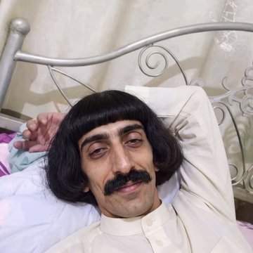 A man wearing a black bob wig and mustache rests in an ornate metal-framed bed with white pillows and a patterned curtain behind him.