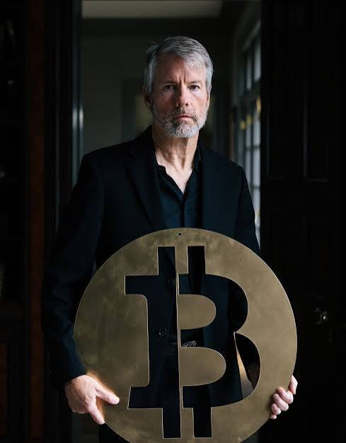 A man in a dark suit holds a large bronze Bitcoin symbol in a dimly lit room.