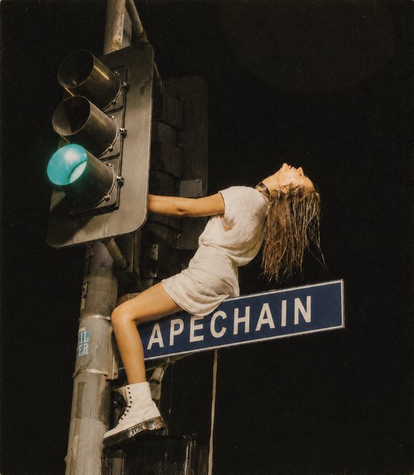 A young woman in a white dress and combat boots is dramatically leaning back against a traffic light pole that has a green light illuminated, with a blue street sign reading "APECHAIN" attached to it. The scene captures the post's promotion of the $APE token on the ApeChain network, using the surreal urban setting to visually represent the brand name.