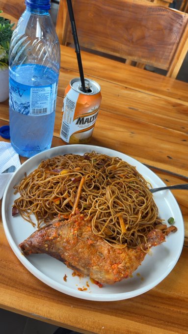 A meal consisting of crispy fried noodles and a red-sauced chicken leg is served on a white plate alongside a bottle of water and a Madrilena soda, capturing the classic fairground cuisine experience.