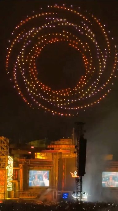 A nighttime outdoor event featuring a spectacular drone light show forming circular patterns in the sky above a stage with 'ARENA' visible and large screens displaying performers.