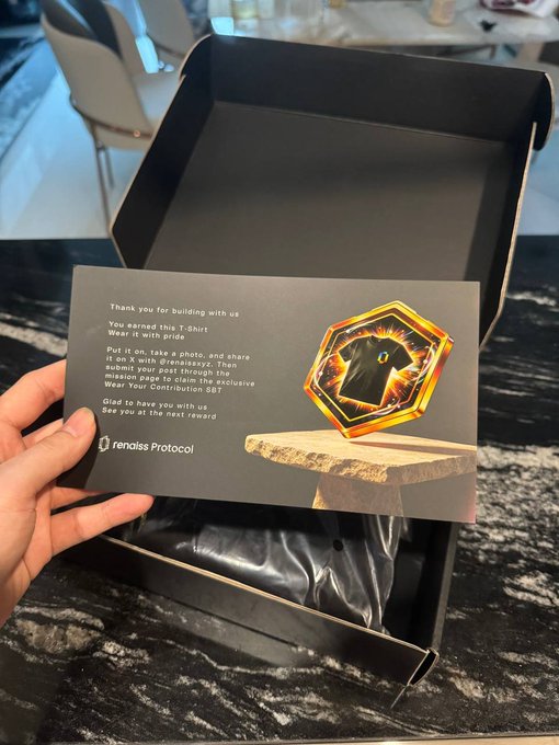 A hand holds a black thank-you card from "renaiss Protocol" that reads "Thank you for building with us. You earned this T-shirt! Wear it with pride" alongside an illustration of a hexagonal golden badge, displayed over an open black merchandise box. The card confirms the tweet-to-claim mechanic for the exclusive "Wear Your Contribution 557" badge mentioned in the post.
