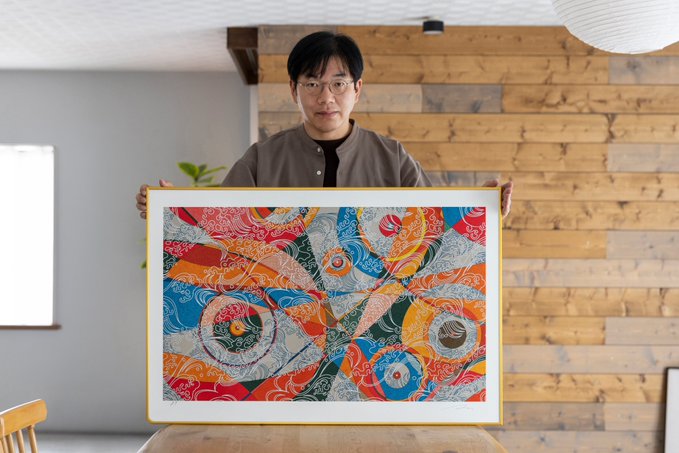 A man in a grey shirt proudly displays a colorful abstract artwork with swirling patterns in front of a wooden accent wall.