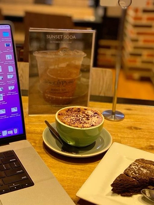 A green bowl of frothy coffee with chocolate sprinkles sits on a wooden table next to a laptop and a sign for 'SUNSET SODA - OCTOBER EXCLUSIVE' at Black Star Pastry, with a slice of chocolate cake nearby.