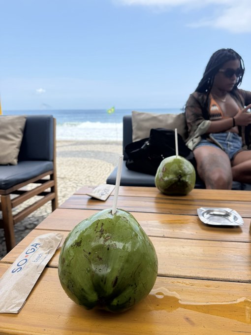 A photograph of two fresh green coconuts with straws on a wooden beachside table, with a woman wearing sunglasses sitting on a lounge chair in the background near the ocean.
