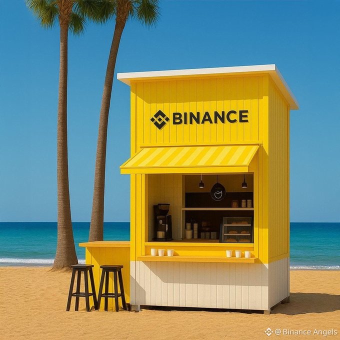 A bright yellow Binance-branded kiosk sits on a sandy beach under palm trees, with the word "BINANCE" prominently displayed on the front. The setup, complete with stools and a small coffee bar area, plays on the post's invite—suggesting a casual "pull-up" for a coffee at Binance's beachside stand. A watermark in the corner reads "@Binance Angels."