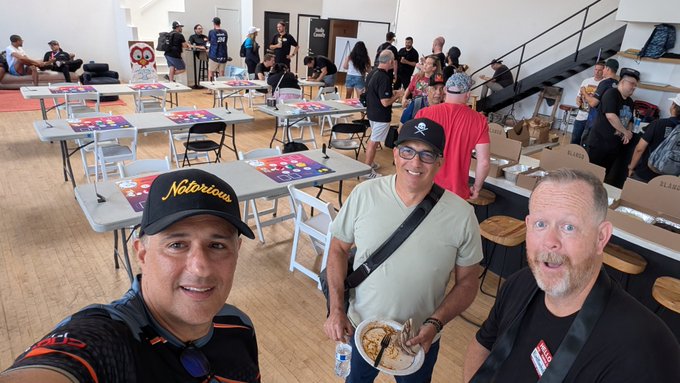Three men pose for a selfie in a bustling event space filled with tables set up for a trading card game tournament, with the man on the left wearing a black "Notorious" cap, the middle man in a white t-shirt and black baseball cap holding a food container, and the right man in a black shirt with an excited expression, while numerous other attendees and game tables stretch into the background of the venue.