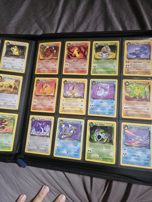 An open Pokémon card binder displaying a collection of holographic cards including Magmar, Charmander, Vulpix, and various other Pokémon, held over a gray bedsheet.