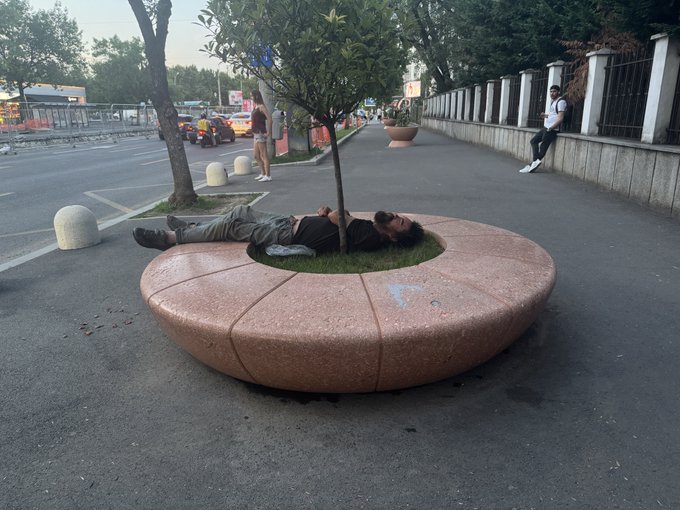 A person is lying on a circular stone bench around a tree on a city sidewalk, while others walk in the background.