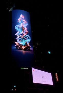 A dim concert venue featuring a tall vertical screen displaying a colorful fantasy character and a lower screen with the name “ELY,” visible from an audience perspective.