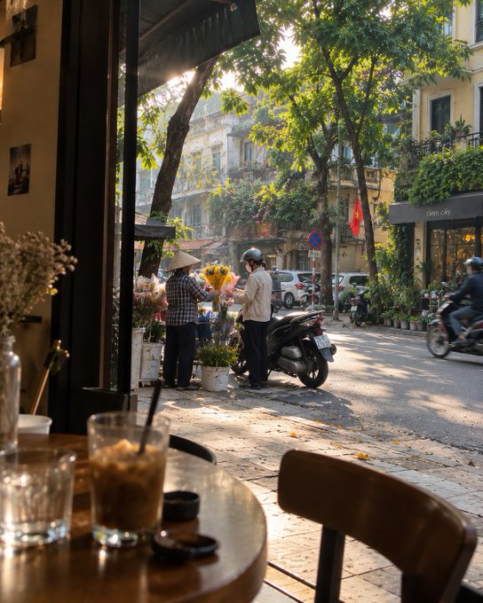 A view from inside a cafe looking out onto a sunny street where a person in a conical hat sells flowers to a customer standing near a parked motorbike.