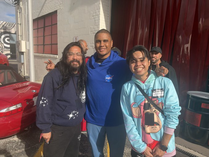 A group of four men pose for a photo outdoors, with a man in a blue NASA hoodie standing centrally between a man in a star-patterned hoodie and a man in a light blue graphic hoodie, with a red sports car visible behind them.