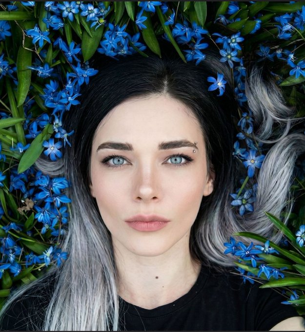 A close-up portrait of a woman with striking blue eyes, dark roots fading into silver-gray hair, wearing a black top, surrounded by vibrant blue flowers and green leaves.