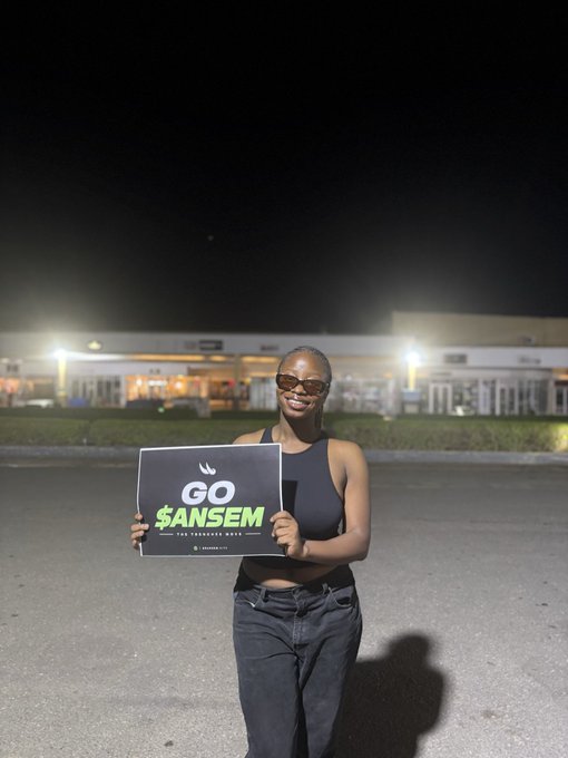 A woman in sunglasses and a black tank top smiles while holding a sign reading 'GO $ANSEM' outside a building at night.
