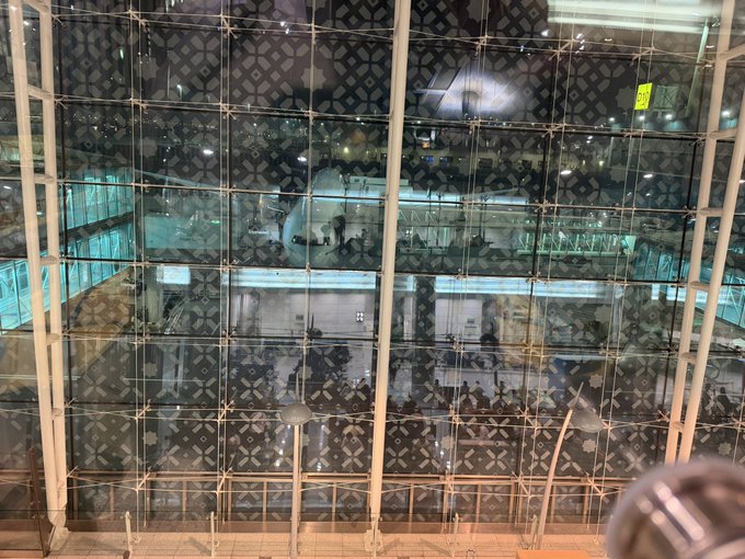 An interior architectural view shows a large glass facade with decorative patterned panels reflecting the surrounding space, with structural beams and a railing visible.