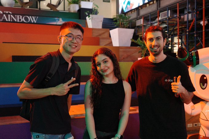 iced tea and a rainbow-colored sign for "Rainbow" are present, with two individuals posing against the backdrop of a large potted plant and a colorful mural.