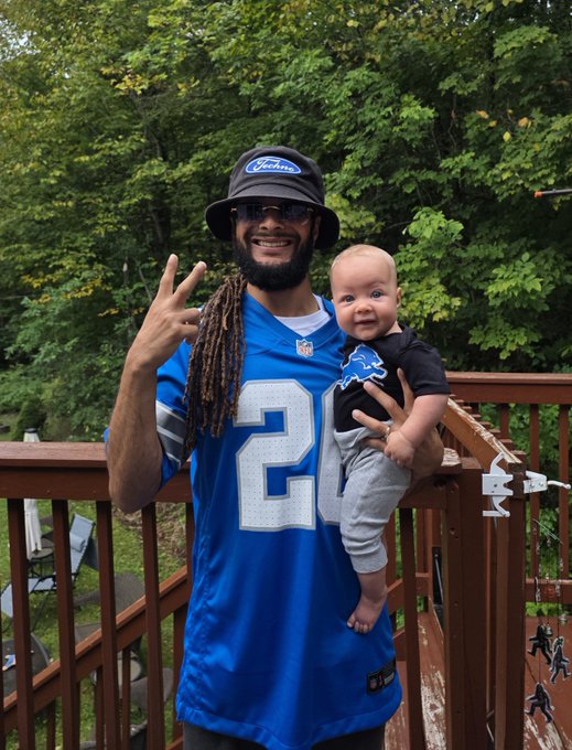 A smiling man wearing a Detroit Lions #20 jersey and a "Tyreek" bucket hat holds a baby on a wooden deck with trees in the background.
