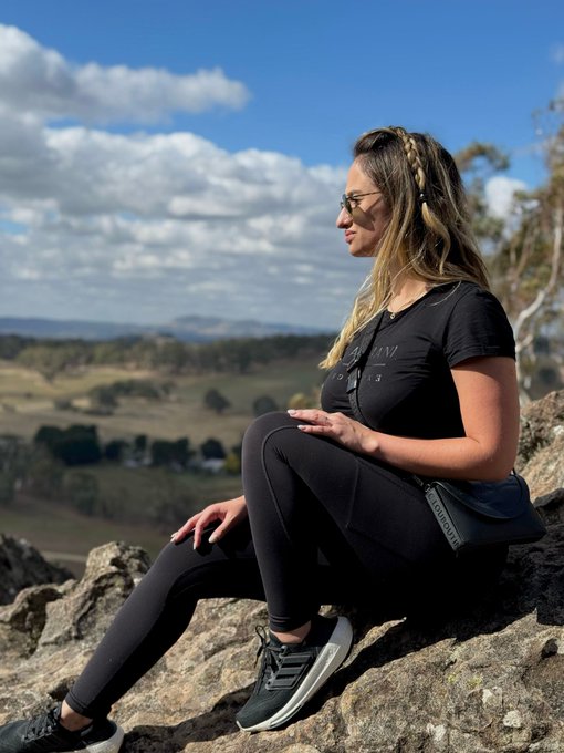A blonde woman wearing sunglasses, a black t-shirt, and black leggings sits on a rocky outcrop overlooking a scenic valley with rolling hills and trees under a partly cloudy sky.