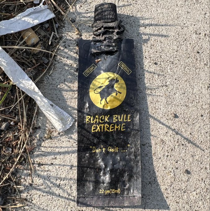 A discarded Black Bull Extreme sachet (22 gm/15ml energy gel) lies on concrete ground with the tagline 'Don't Quit...' visible on its black and gold packaging.