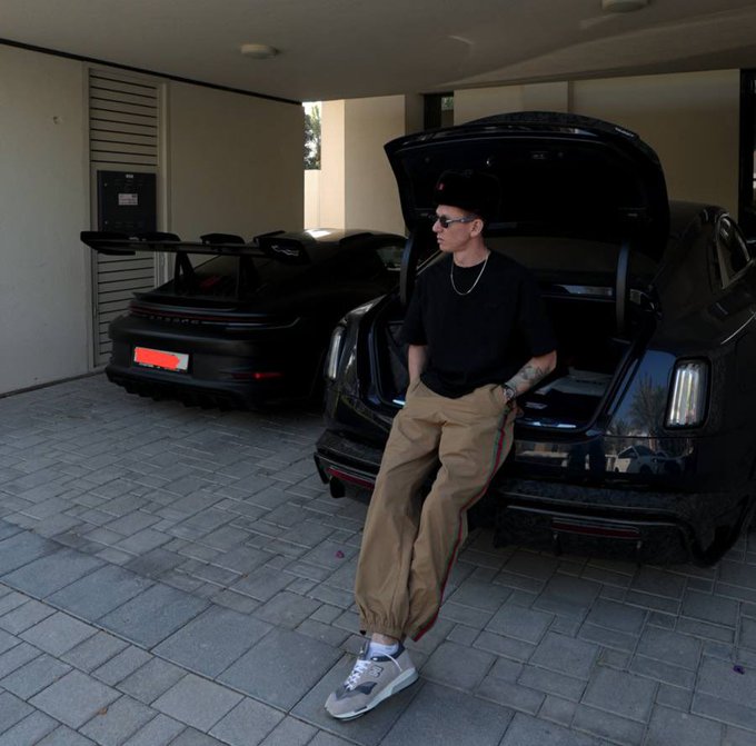 A man in a black t-shirt, beige track pants with red and green stripes, a black cap, and gray sneakers sits casually against the open trunk of a black Rolls-Royce Cullinan in a garage, with a black Porsche 911 GT3 RS parked behind him. The image aligns with the post's theme of learning to trust one's own judgment over outside opinions, visually conveying the kind of quiet self-reliance and personal success that comes from finding one's own footing after a difficult period.