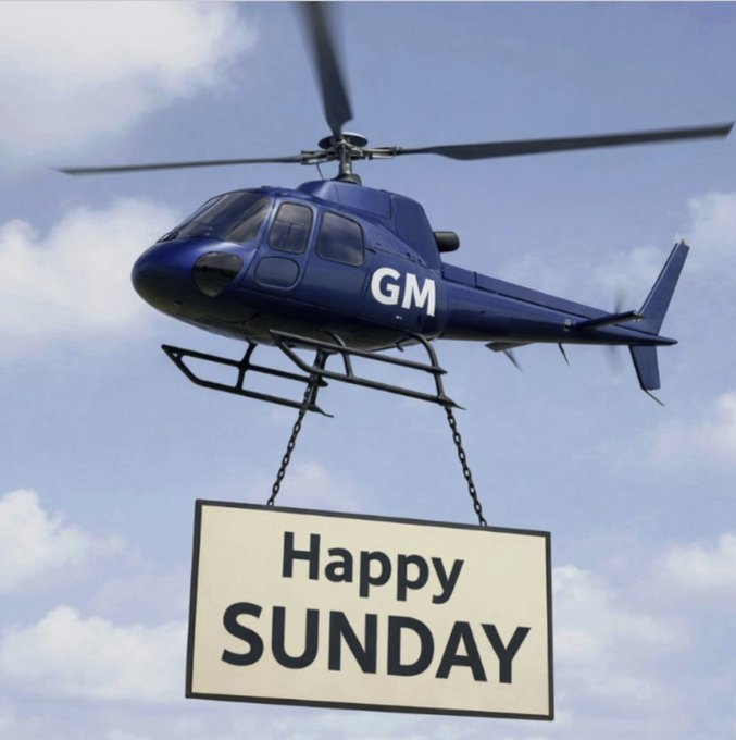 A blue helicopter with "GM" on its side flies through a cloudy sky, carrying a sign that reads "Happy SUNDAY" suspended by chains.