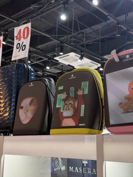 Backpacks featuring pixel-art NFT characters from Chimpers and Cool Cats are displayed on a retail shelf at a Spanish airport, with a "40%" discount sign hanging above. The "MASER" text partially visible at the bottom suggests a Maserati-branded display nearby.