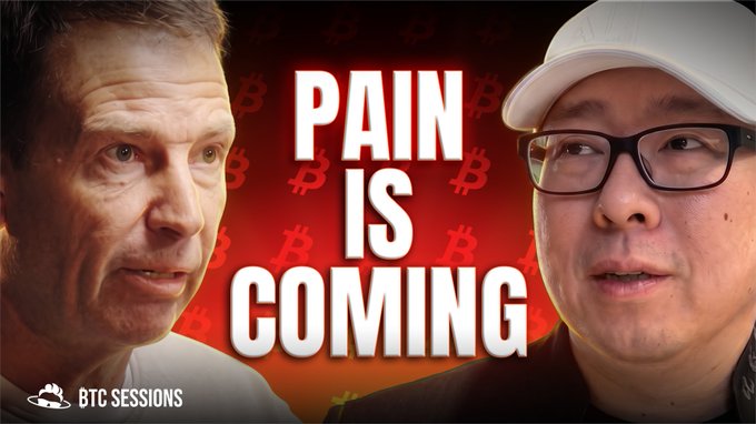 The image features a man wearing glasses with a hat, standing next to another person who also has glasses and a hat on. The background of the image is red, which appears to be part of an advertisement or promotional material for a product or service related to Bitcoin. The text "Pain is coming" is written in white above the two individuals' heads, possibly indicating that they are experiencing some sort of discomfort or pain.