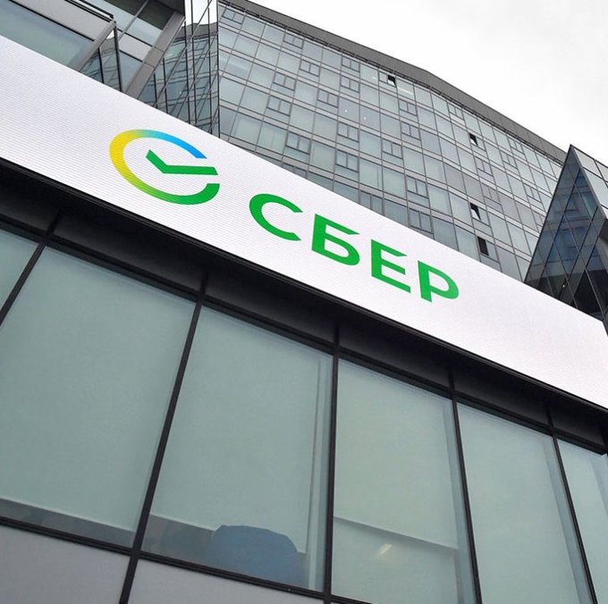 A low-angle photograph shows the exterior of a modern glass office building featuring a large illuminated sign with Sberbank's green and yellow "G"-shaped logo and the Cyrillic text "СБЕР" (Sber), Russia's largest bank that is now accepting Tether USDT as loan collateral for institutional clients.